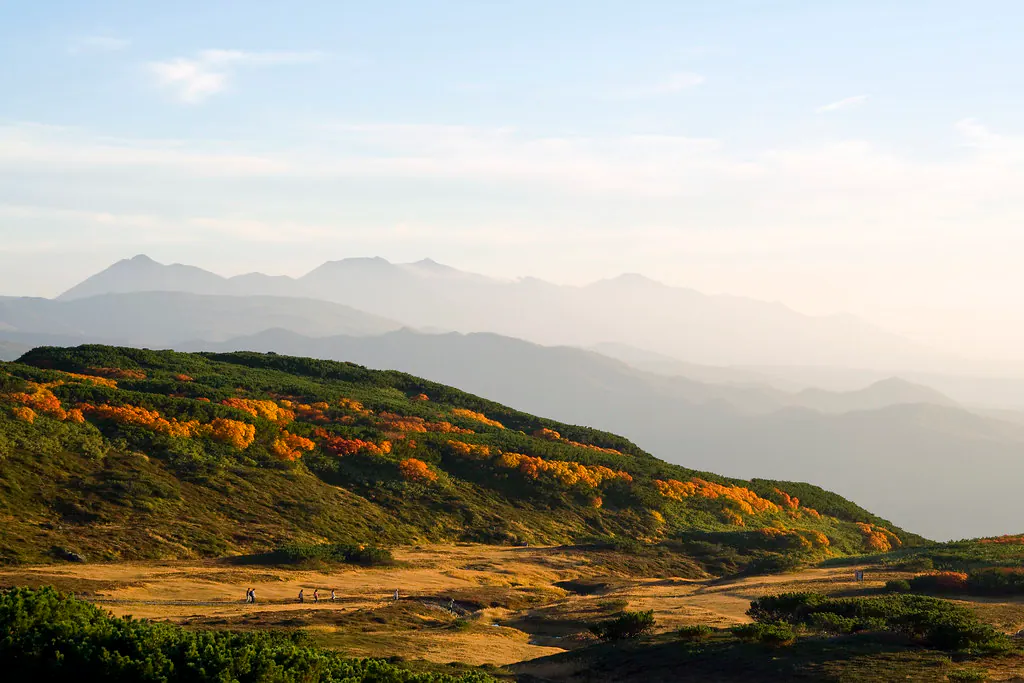 Orange and yellow foliage across open mountain slopes in Daisetsuzan, with walkers on a path and layered peaks in the distance